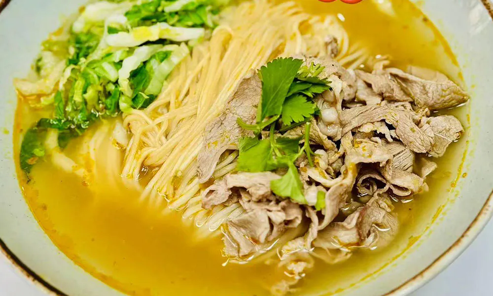 Ceramic bowl of noodle soup in golden spicy broth, loaded with sliced lamb, fresh cilantro and chopped cabbage. Overlaid text: Golden spicy Lamb noodle soup with red chili emojis.