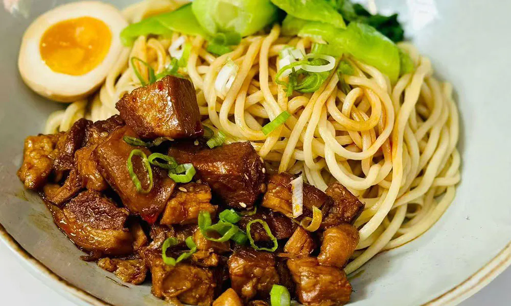 A wide ceramic bowl holds dry noodles topped with glossy braised pork cubes, halved soft-boiled egg, green bok choy and chopped green onions. Text reads Braised pork Dry noodles at bottom.